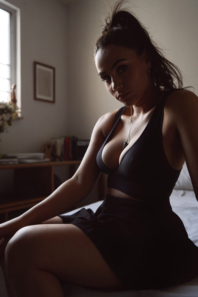 Valeria Cisneros poses in a room with soft lighting wearing a black top and skirt with a necklace and earrings and a vase with flowers in the background.